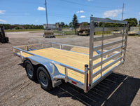 A tandem axle Silver Ox aluminum utility trailer with a silver body, wooden floor, and metal frame, equipped with a ramp gate, parked on a concrete ground with a blue sky in the background.