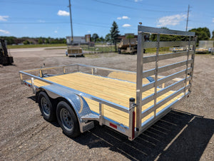 A tandem axle Silver Ox aluminum utility trailer with a silver body, wooden floor, and metal frame, equipped with a ramp gate, parked on a concrete ground with a blue sky in the background.