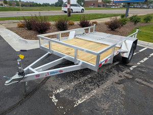 Silver Ox utility trailer parked in a parking lot with a building and cars in the background.