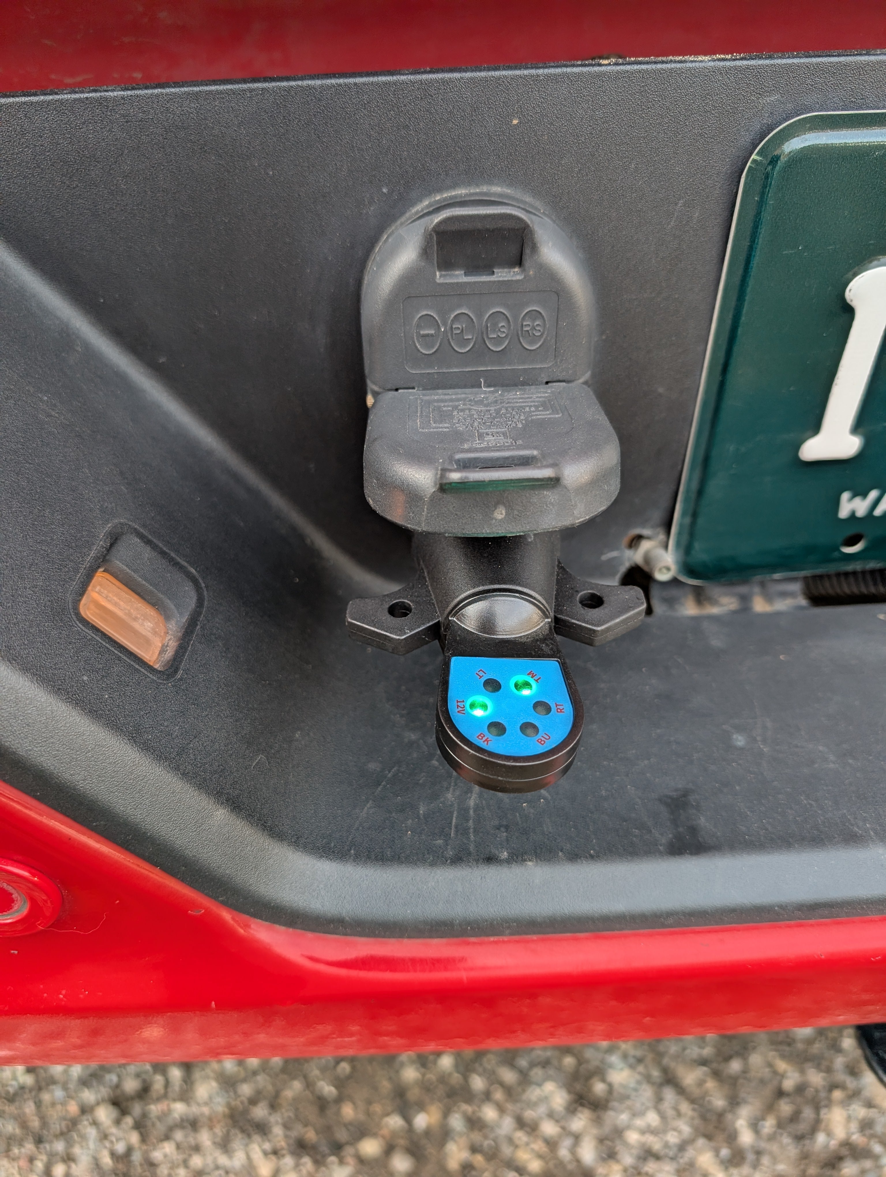 Close-up of a 7 way trailer circuit tester receiver on a vehicle, with a green license plate partially visible.